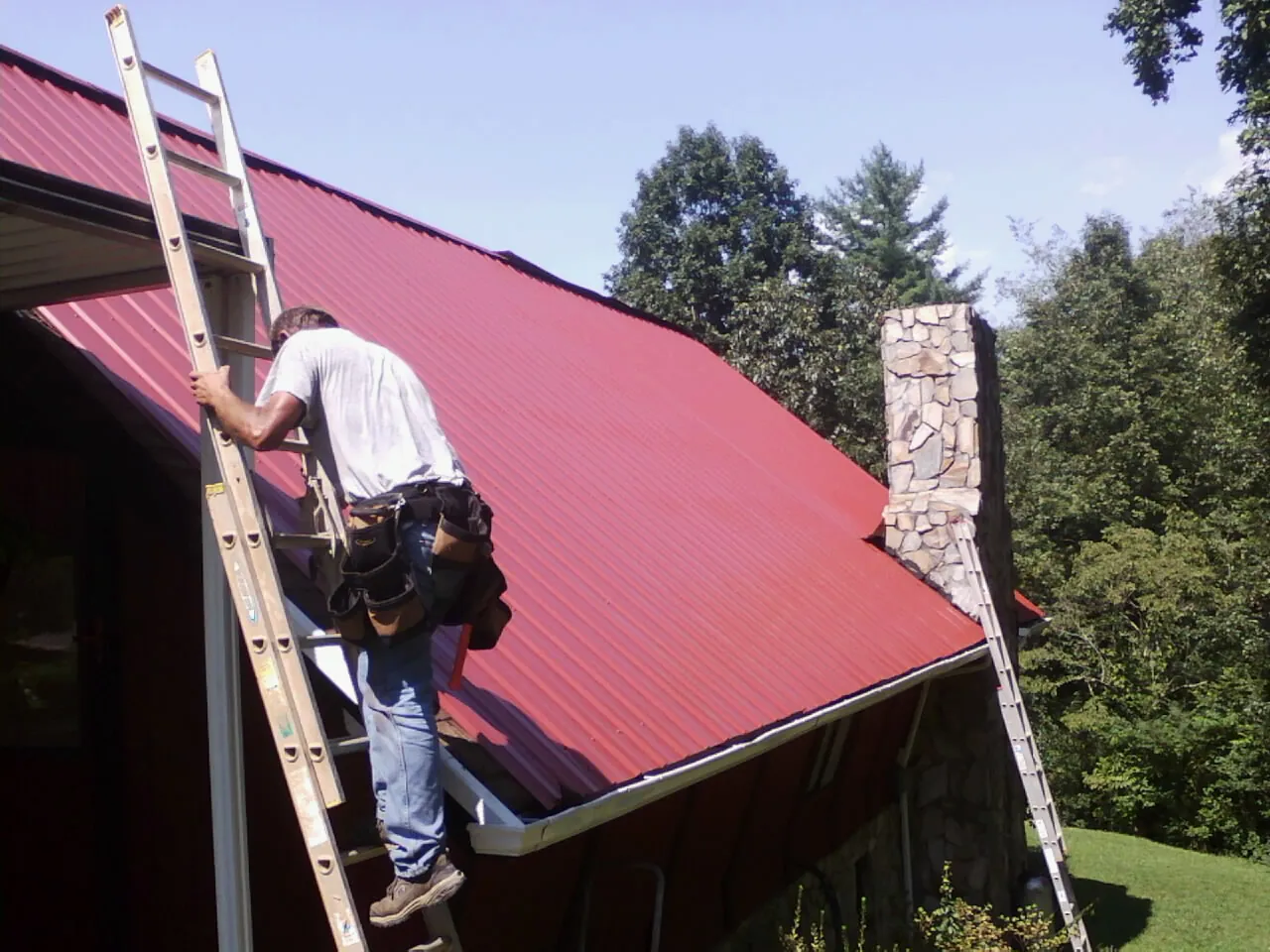 Roofer on a ladder beside a red metal roof and stone chimney — Soffit & Fascia Repair by Roofing Brothers in Lock Haven